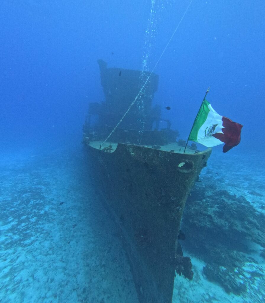 Scuba diver ascending next to the flag-adorned bow of the C-56 wreck on a 'Triton Diving' excursion in Cozumel.